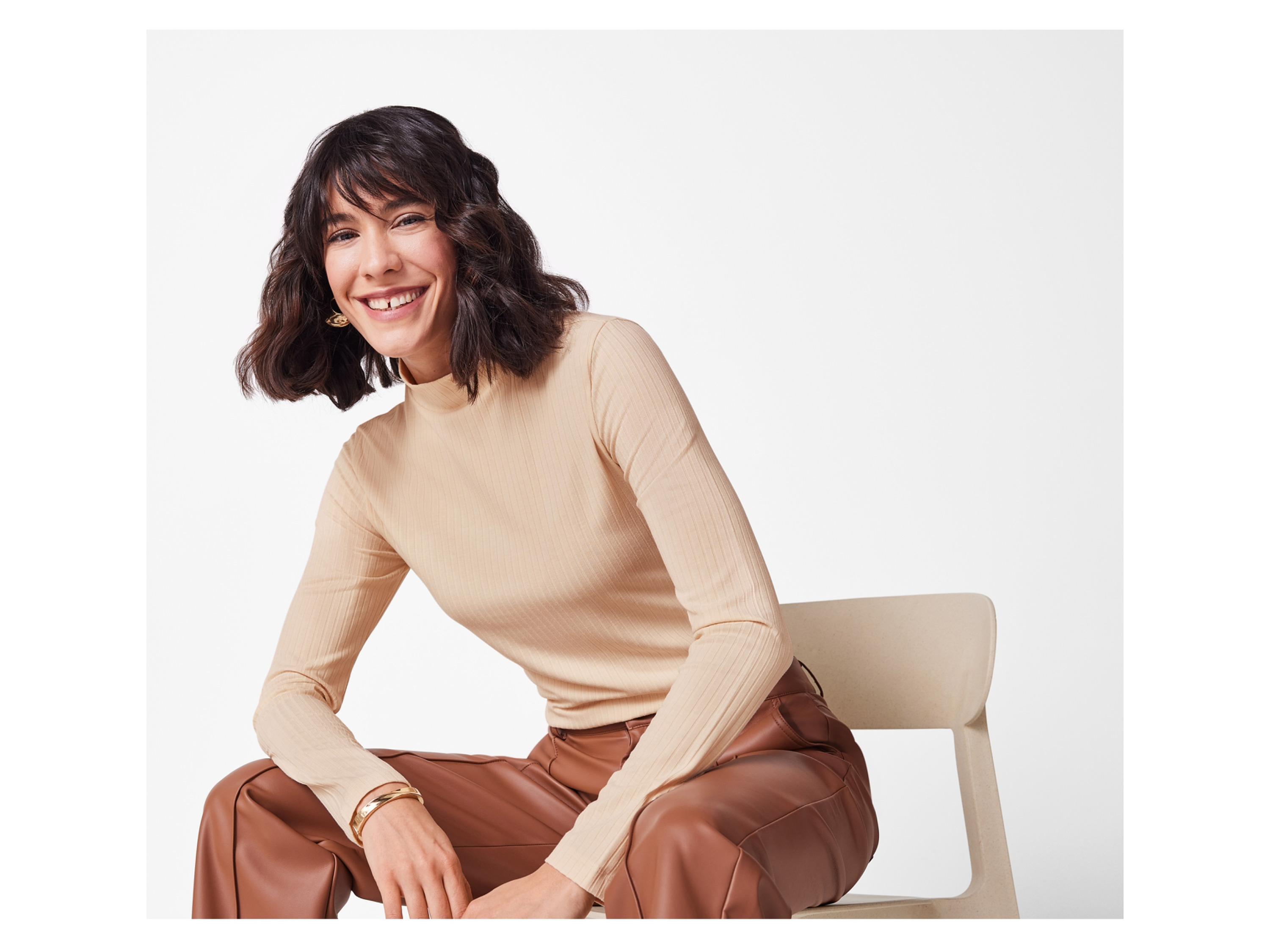 A woman in a beige ribbed turtleneck and brown faux leather pants sits on a chair, smiling.