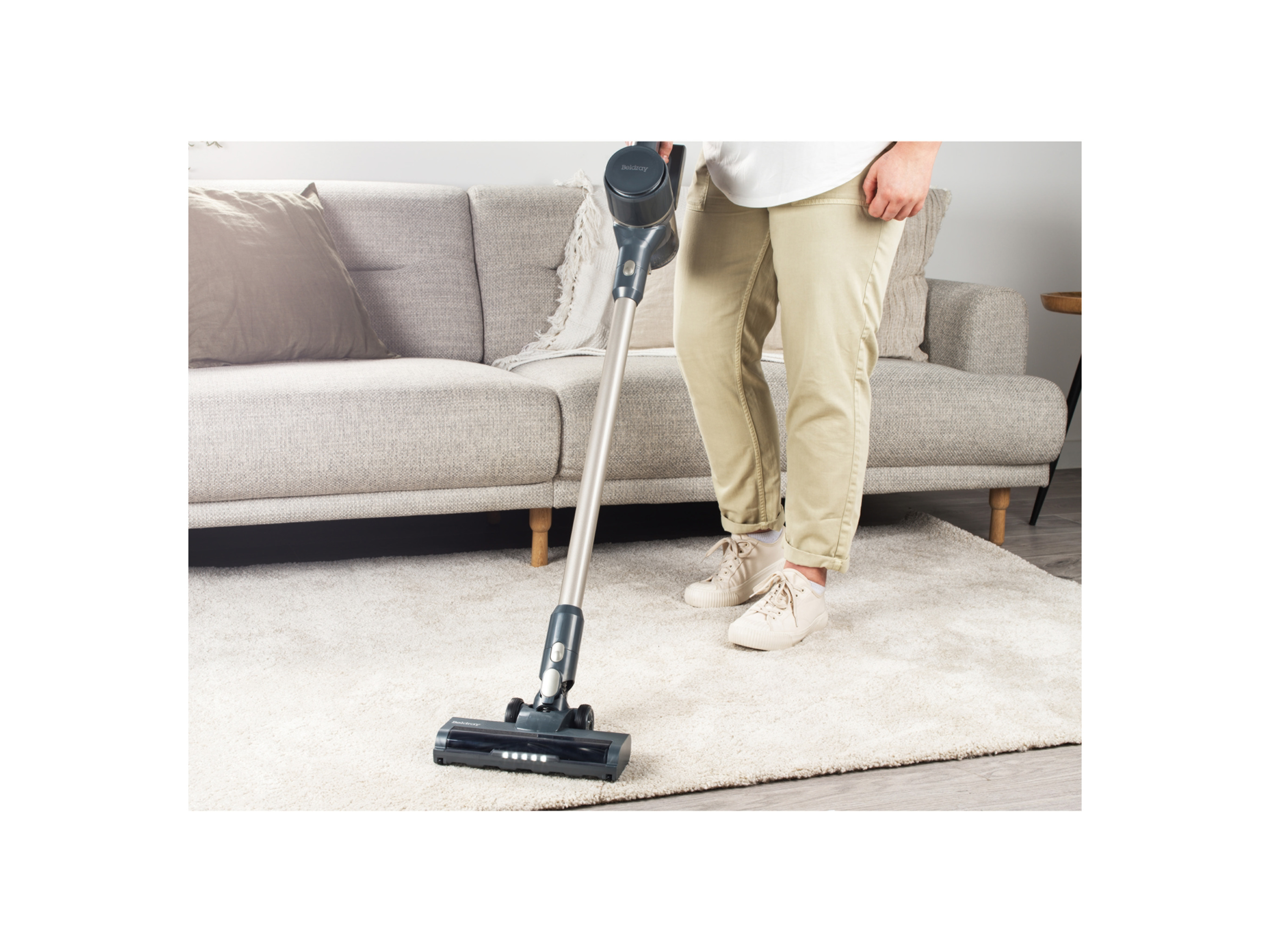 A person uses a Belray cordless stick vacuum to clean a light beige rug in a living room.