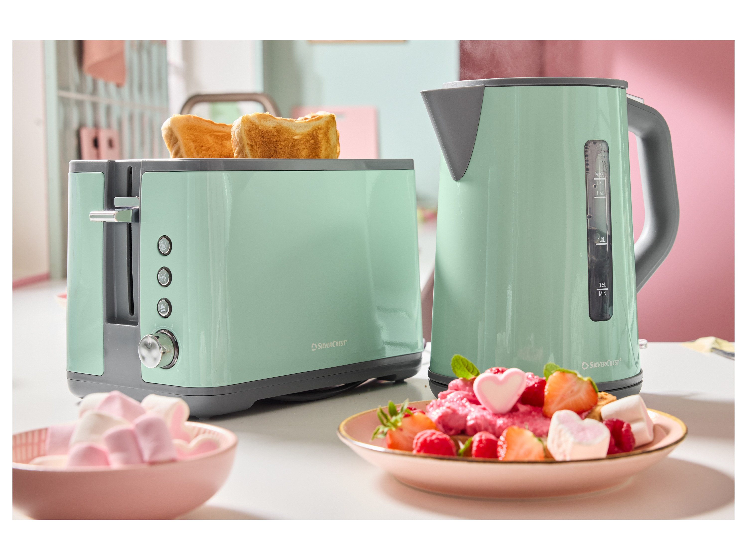 A mint green toaster with toast and a matching kettle next to a bowl of fruit and marshmallows.