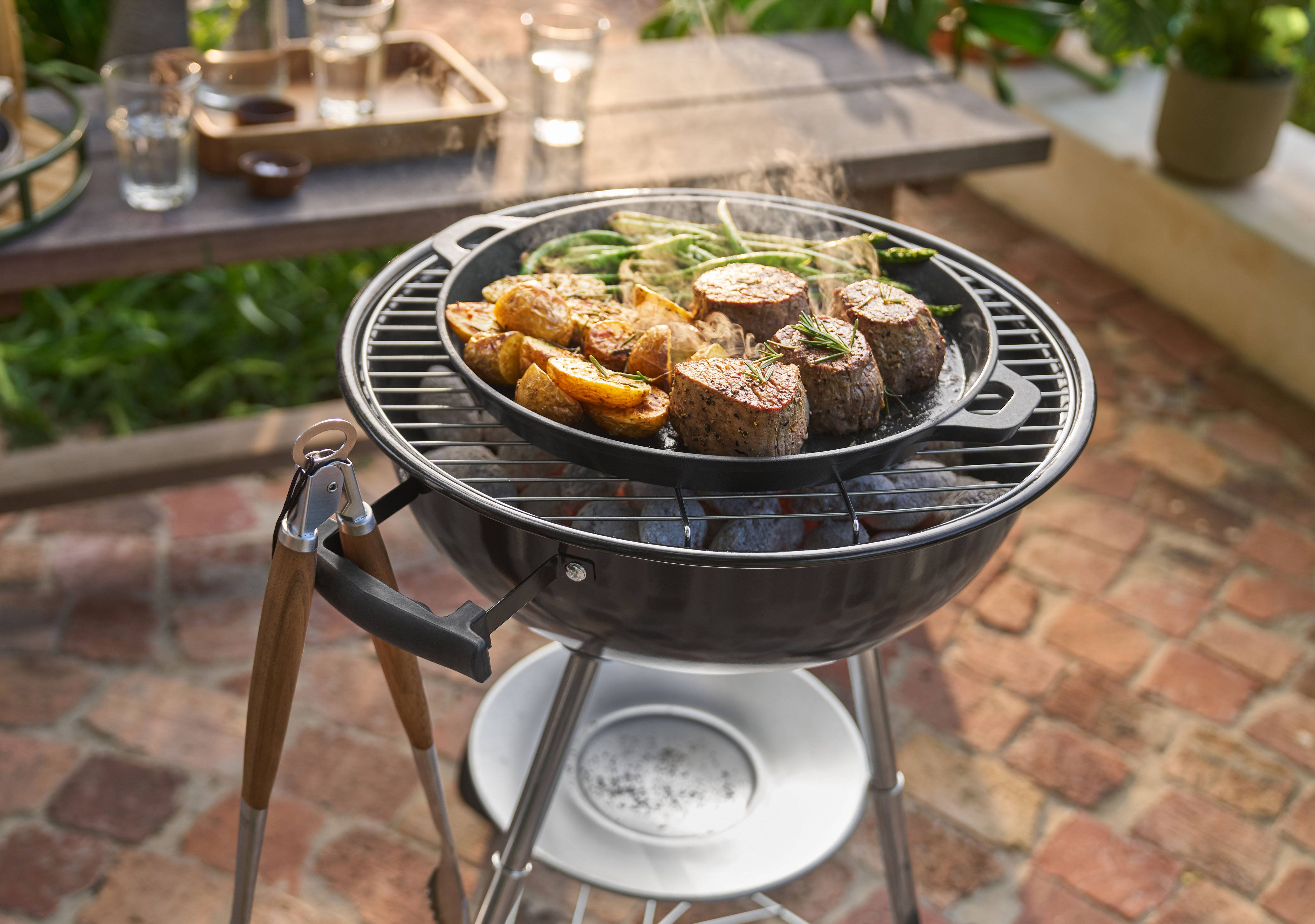 Barbecue with meat, potatoes, and green beans cooking in a pan outdoors.