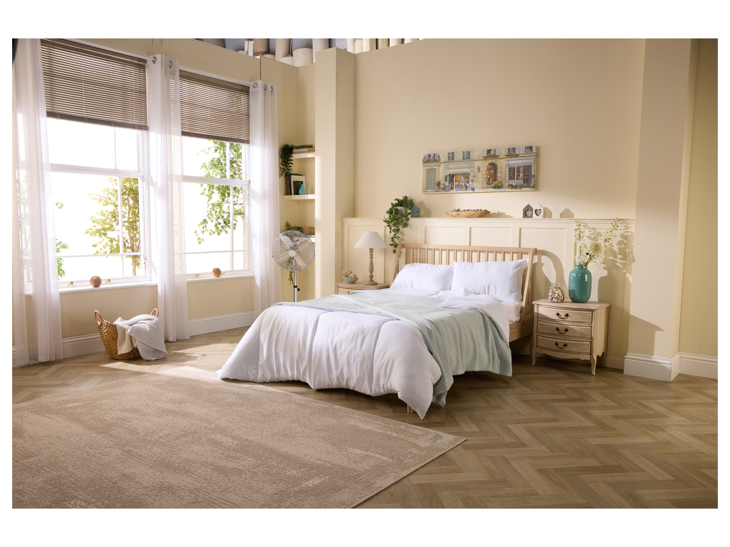 A light and airy bedroom features a wooden bed, a beige rug, and a standing fan.