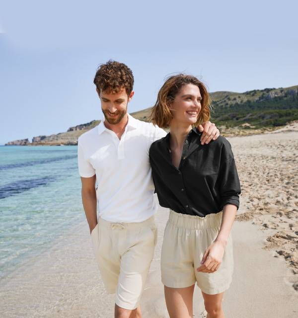 Couple walking on a beach, man in white polo shirt and beige shorts, woman in black shirt and beige shorts.