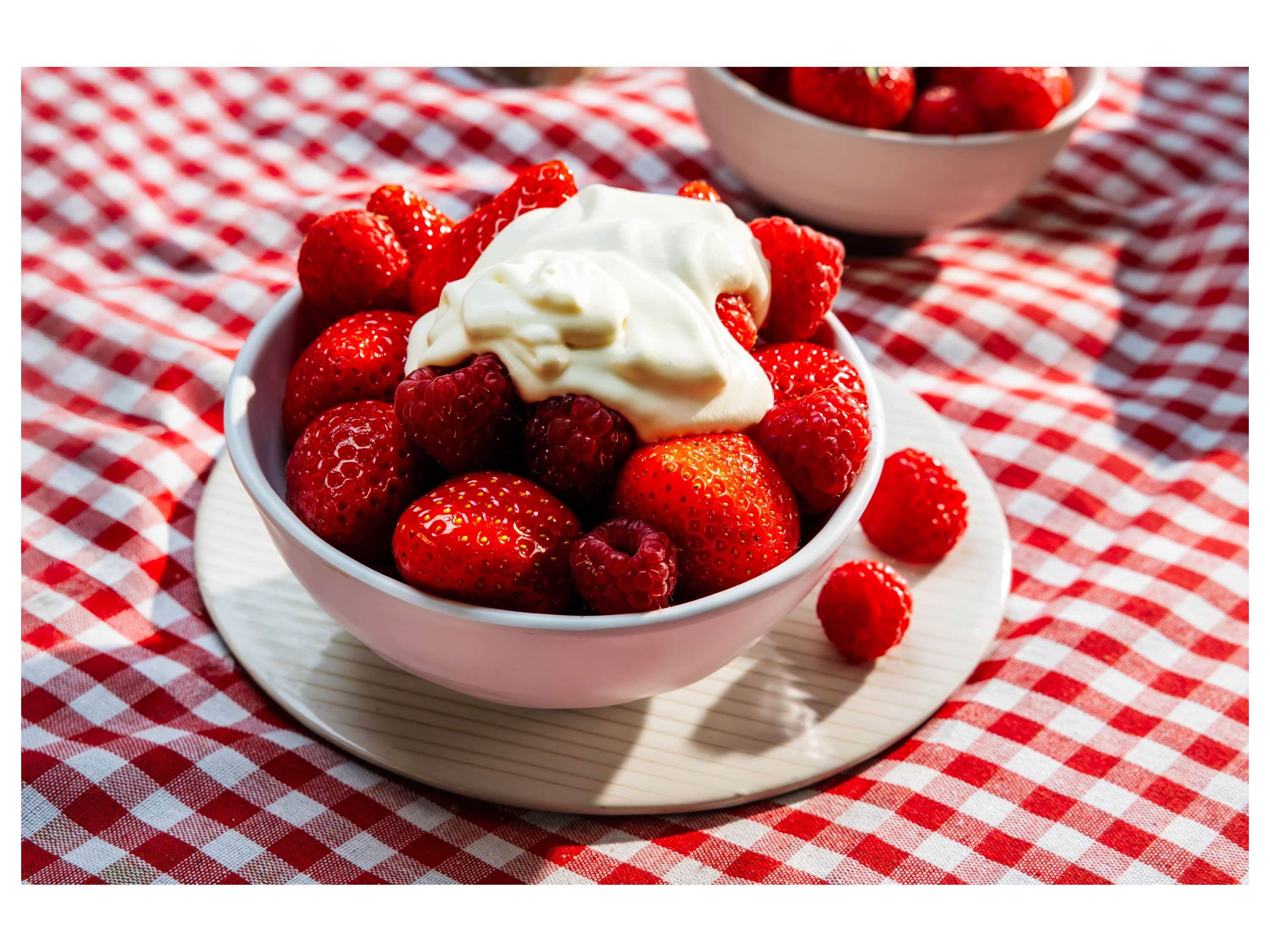 A bowl of fresh strawberries and raspberries topped with whipped cream sits on a red and white checkered tablecloth.