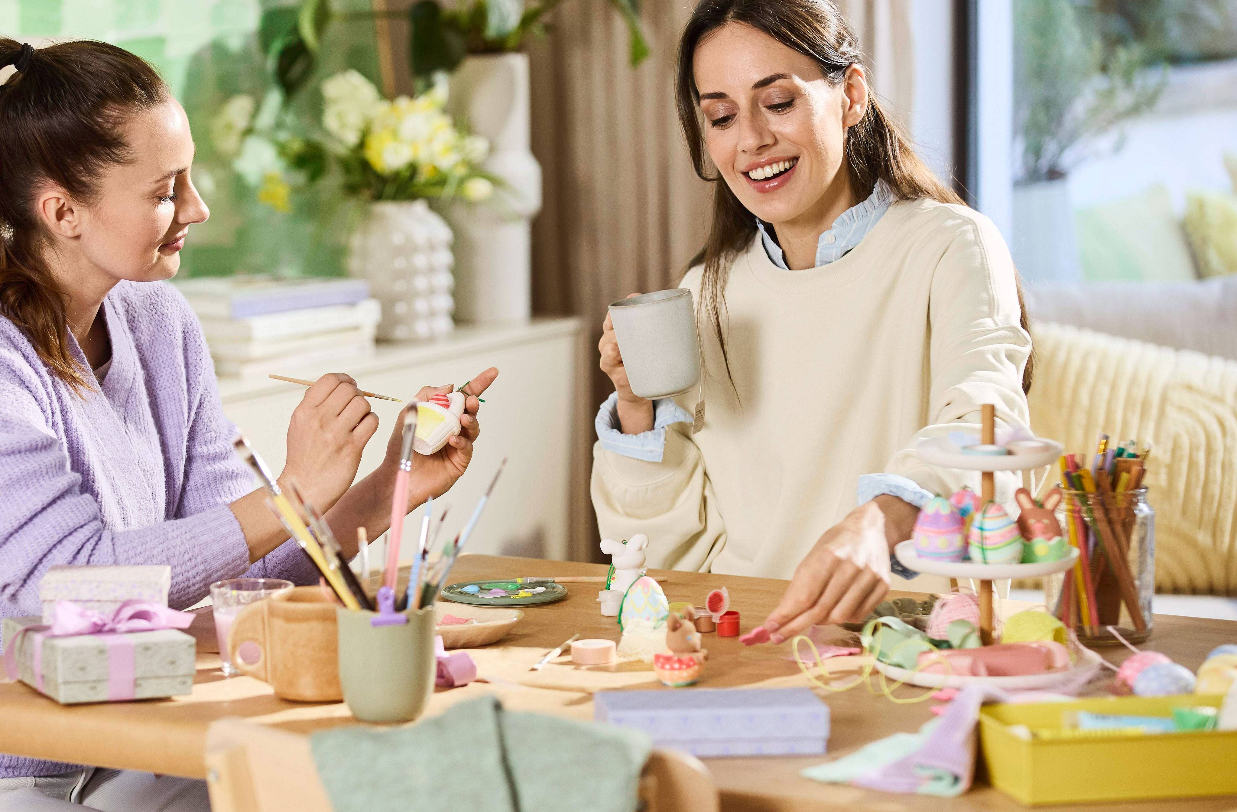 Two women painting Easter eggs and decorations, table full of art supplies.