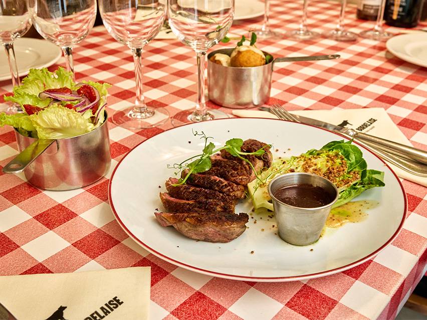 Sliced steak with salad, sauce, and sides on a red and white checkered tablecloth.
