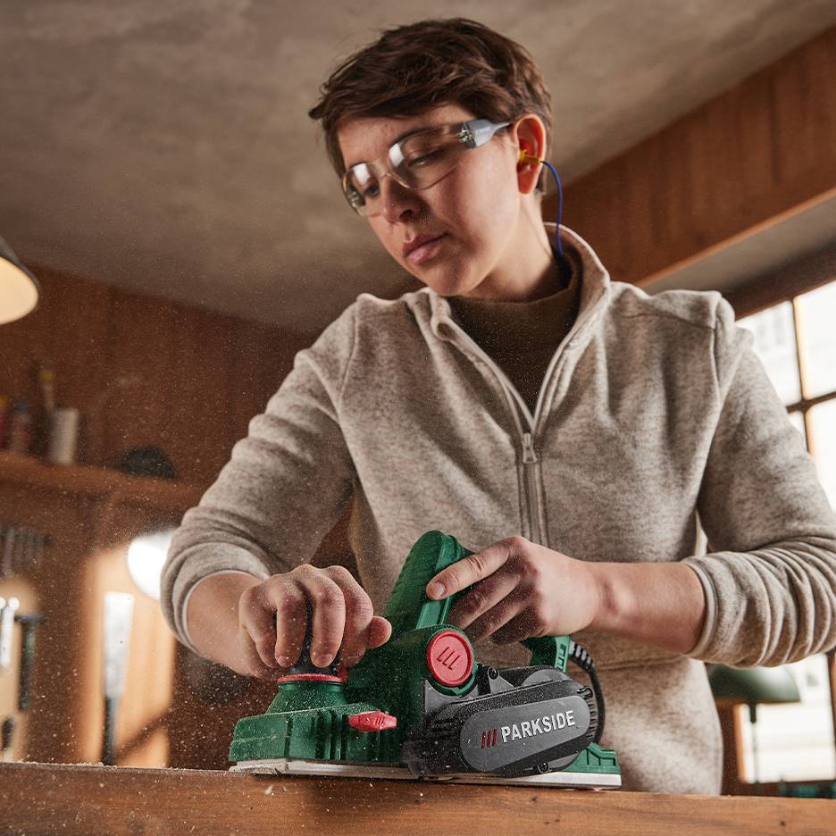 Woman planing wood with a Parkside electric planer, with sawdust flying around.