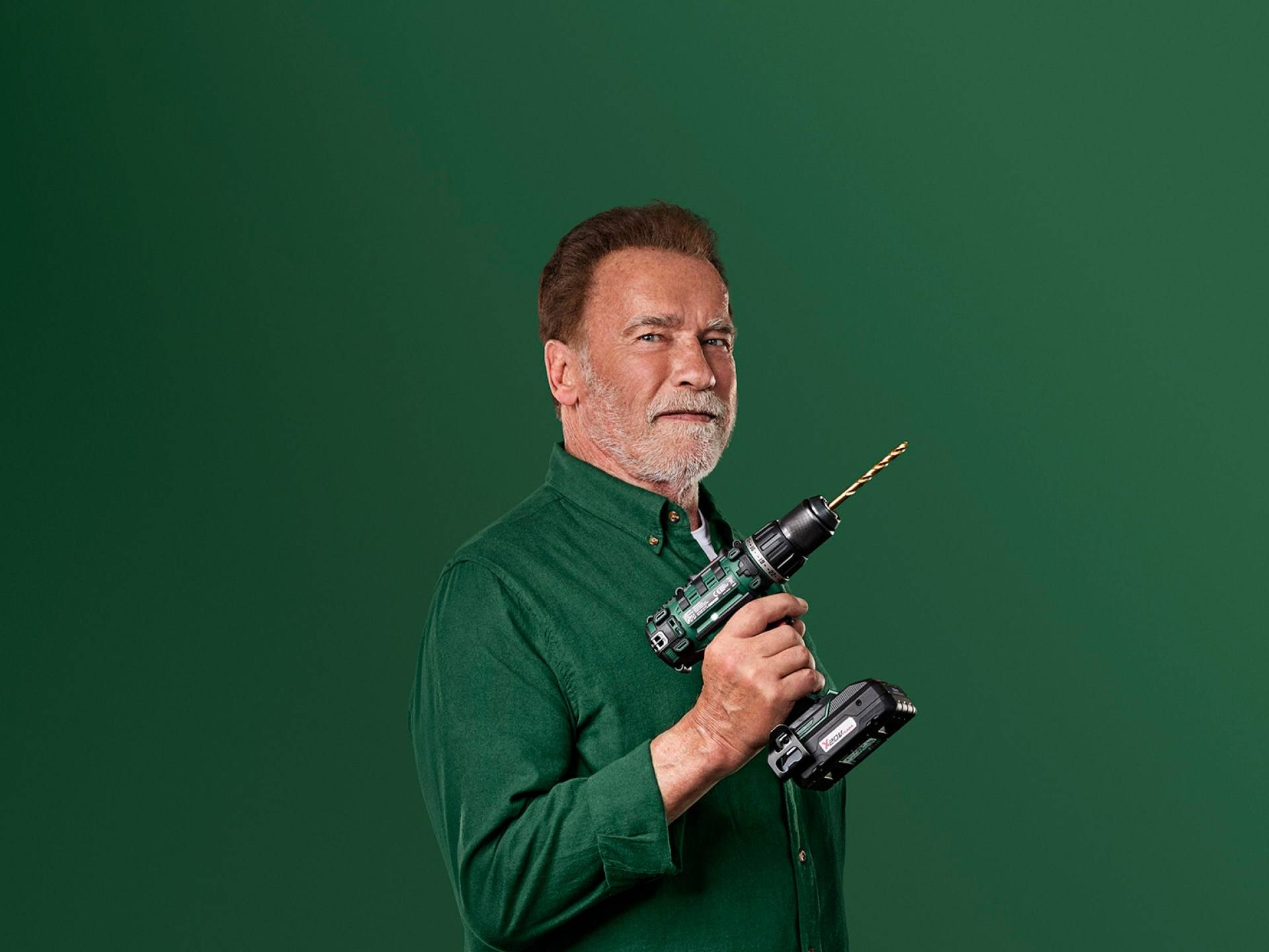 Arnold Schwarzenegger holds a green electric drill against a green background.