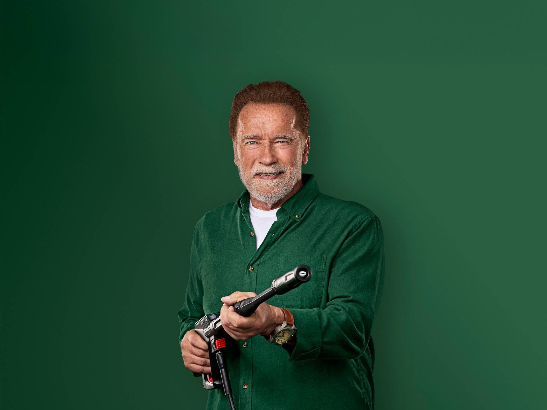 Arnold Schwarzenegger smiles, holding a pressure washer gun.