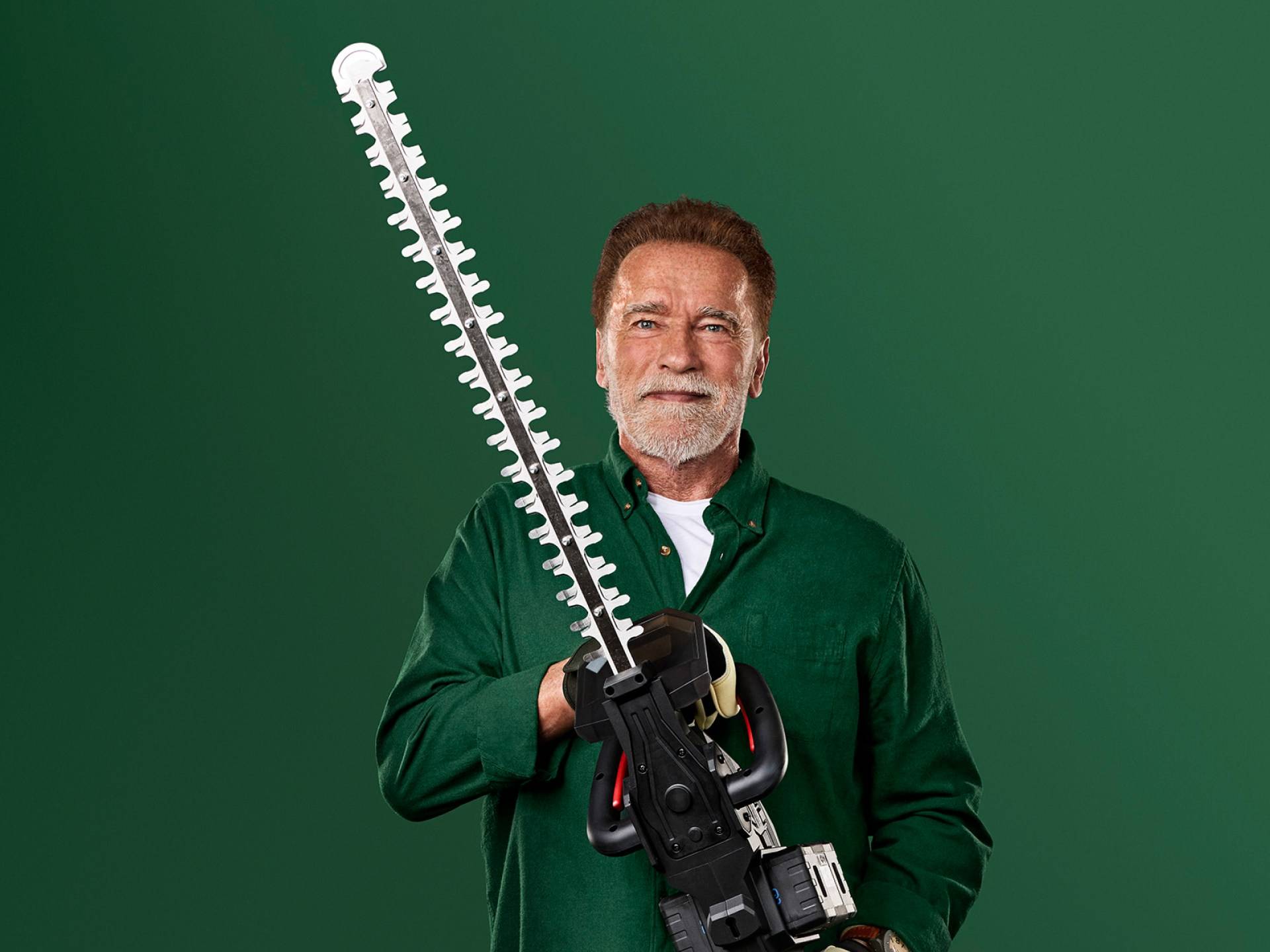 Arnold Schwarzenegger holds a large hedge trimmer against a green background.