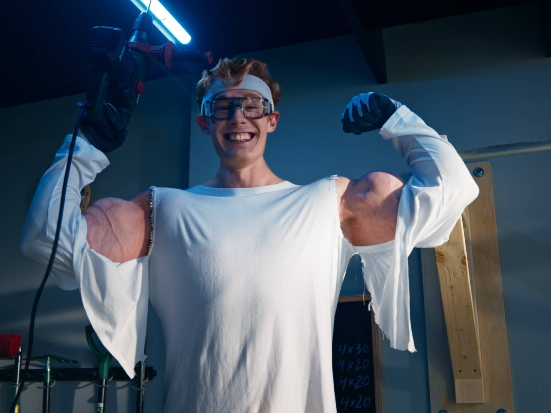 Man with tools and oversized muscles smiling in a workshop.