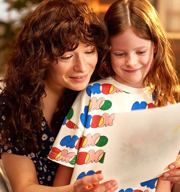 Mother and daughter smiling, looking at a drawing. Daughter wears a colorful t-shirt.