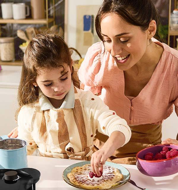 Mother and daughter decorating a waffle with raspberries and cream, with a waffle maker nearby.