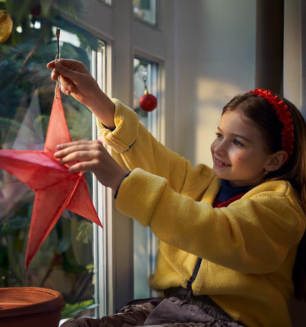 A smiling girl in a yellow cardigan hanging a red paper star decoration on a window.