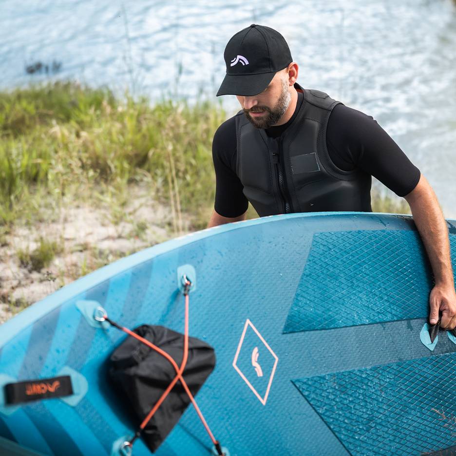 Man in life vest and cap holding a blue paddleboard by the water.