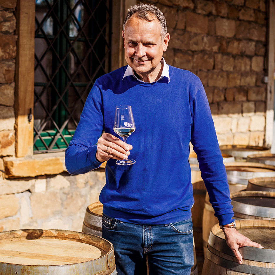 Smiling man in blue sweater holding a wine glass, with wooden barrels in the background.