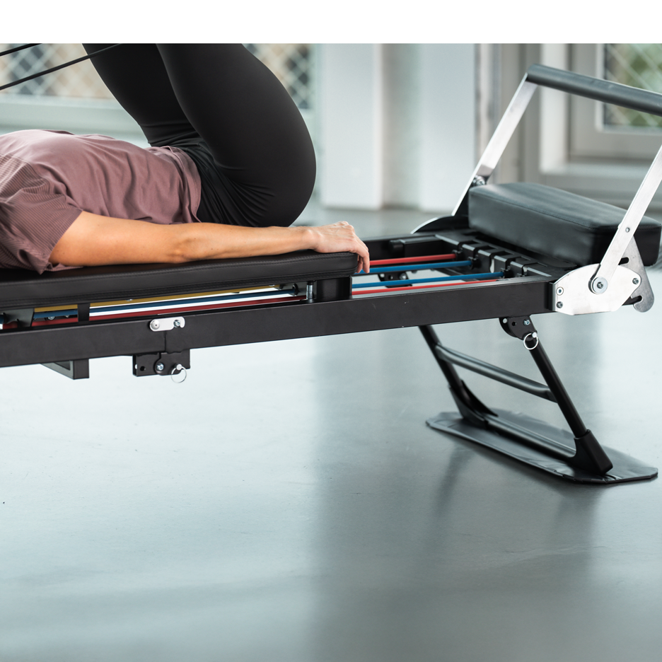 Woman exercising on a black Pilates machine with colored resistance bands.