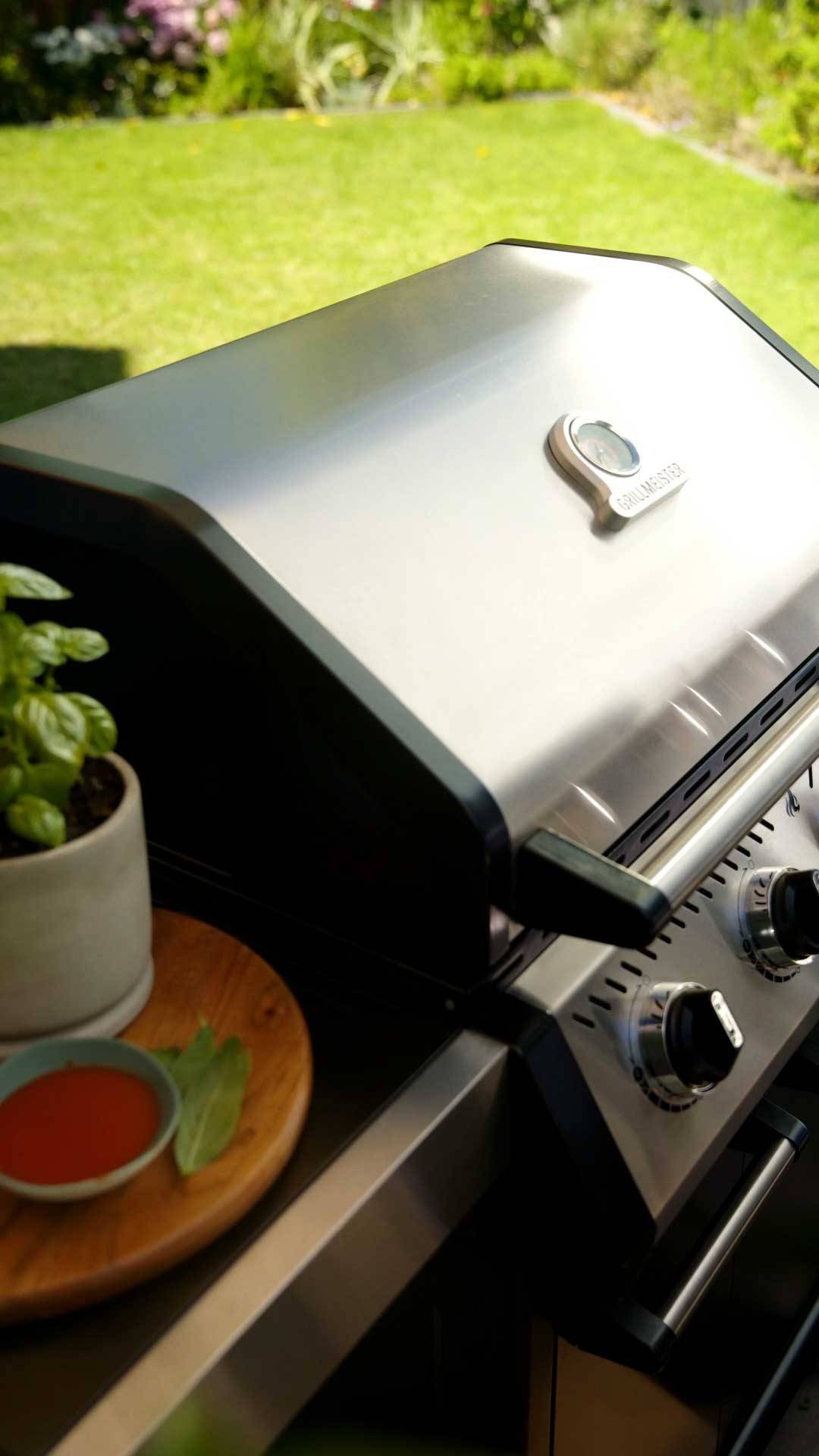 Stainless steel gas barbecue with thermometer, next to a basil pot and sauce.