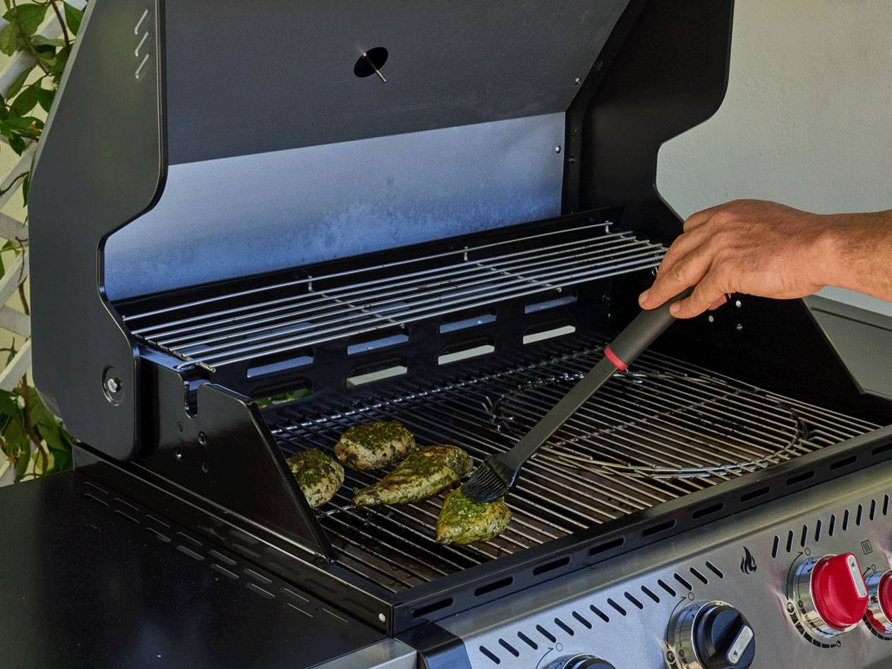 A person basting seasoned chicken pieces on a gas grill with a basting brush.