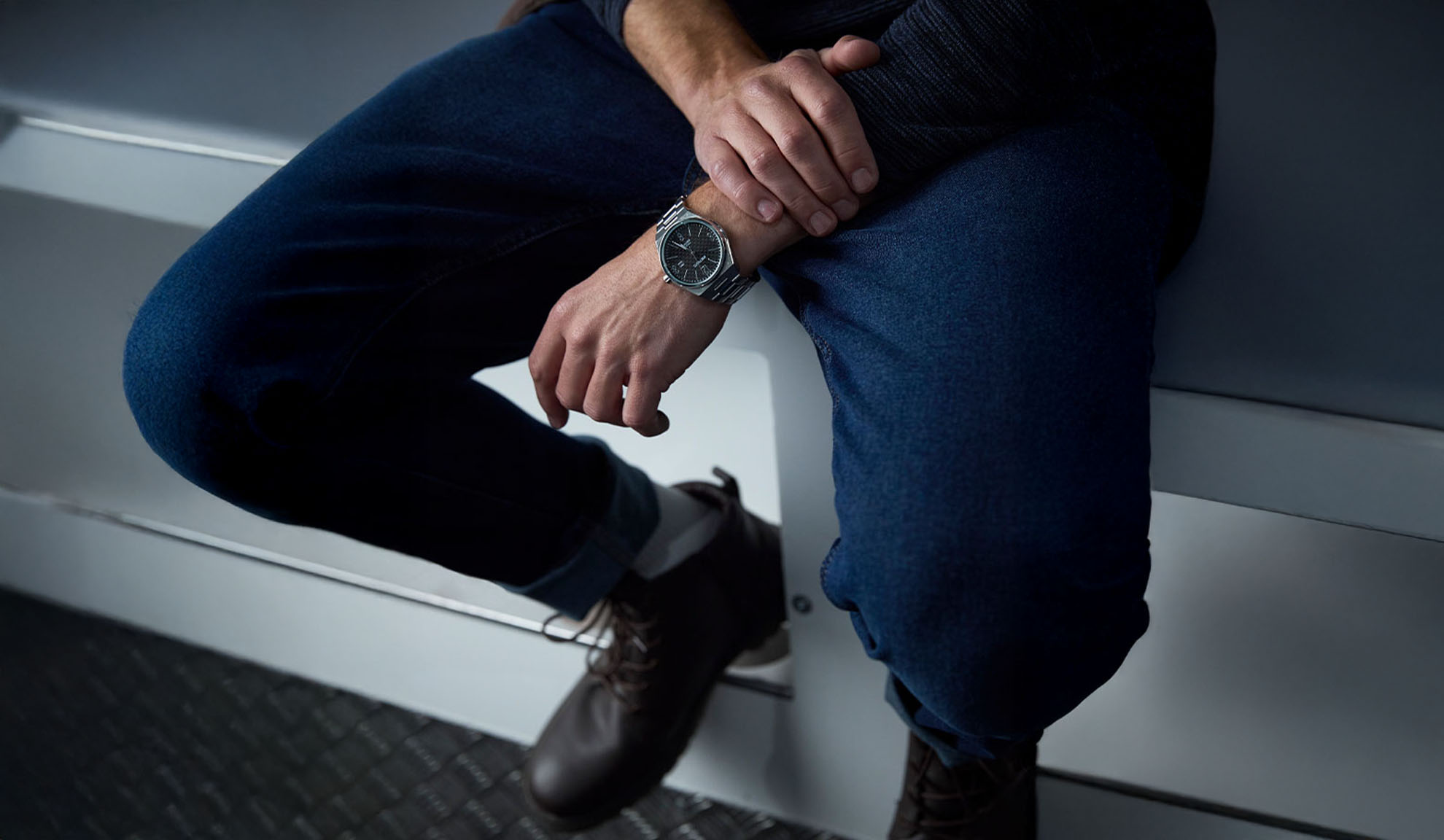 Man wearing dark blue jeans, a dark sweater, and a silver watch, sitting down.