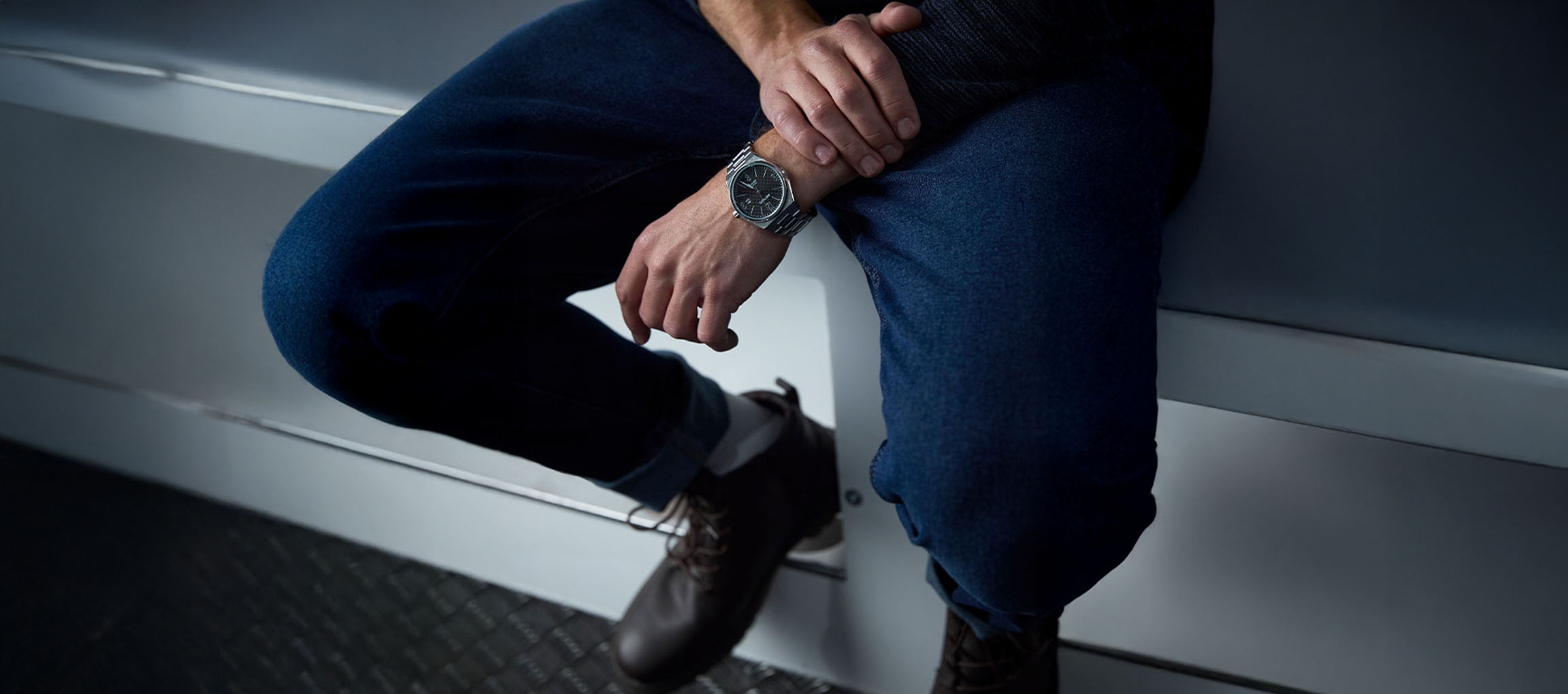 Man wearing blue jeans, brown boots, and a silver watch, sitting on a white surface.