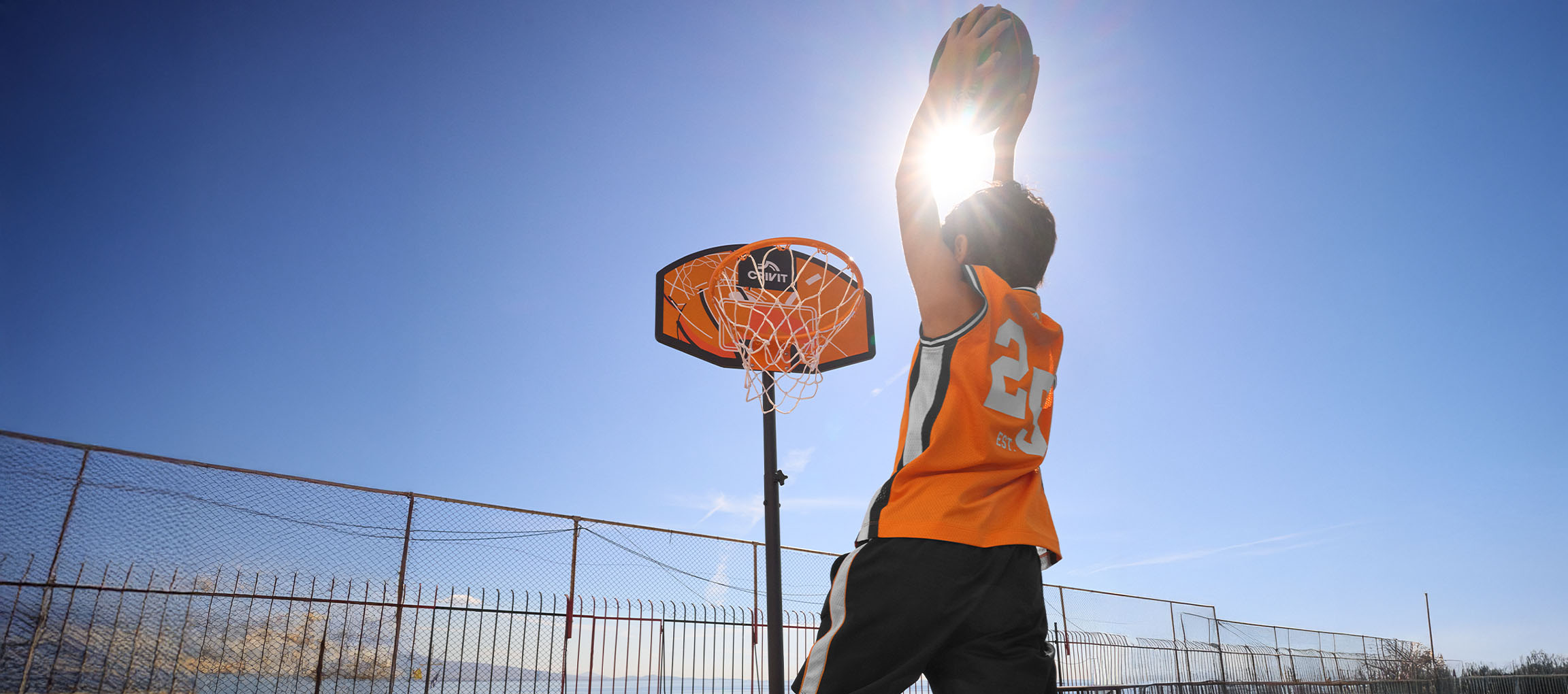 Boy in an orange jersey shooting a basketball against a blue sky.