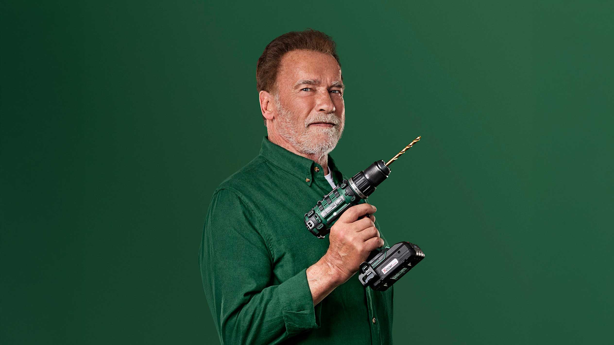 Arnold Schwarzenegger holding a green cordless drill with a gold drill bit.