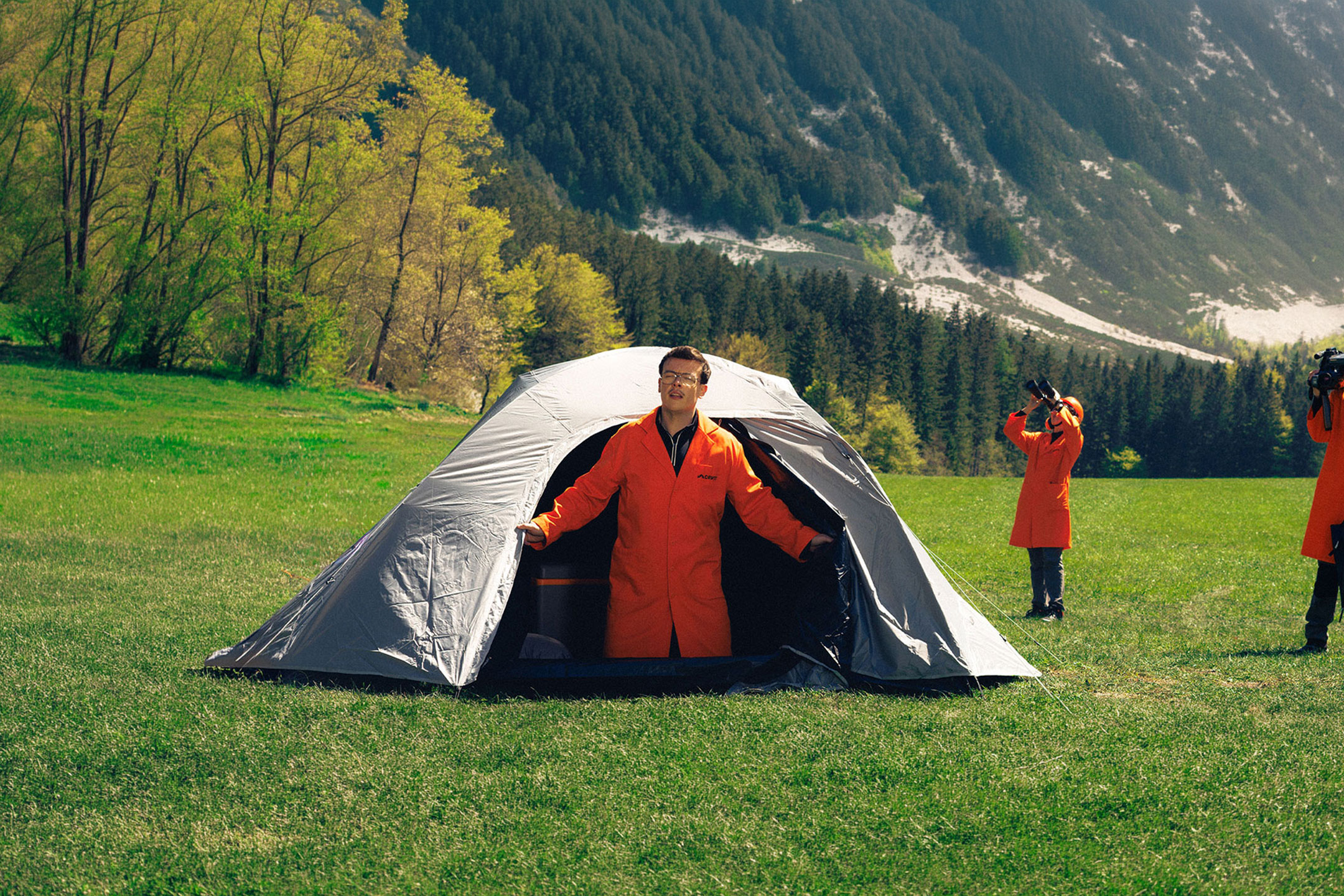 aiLabeling.aiLabelForImageAltText: Three people in orange lab coats with camping and observation equipment in a meadow.