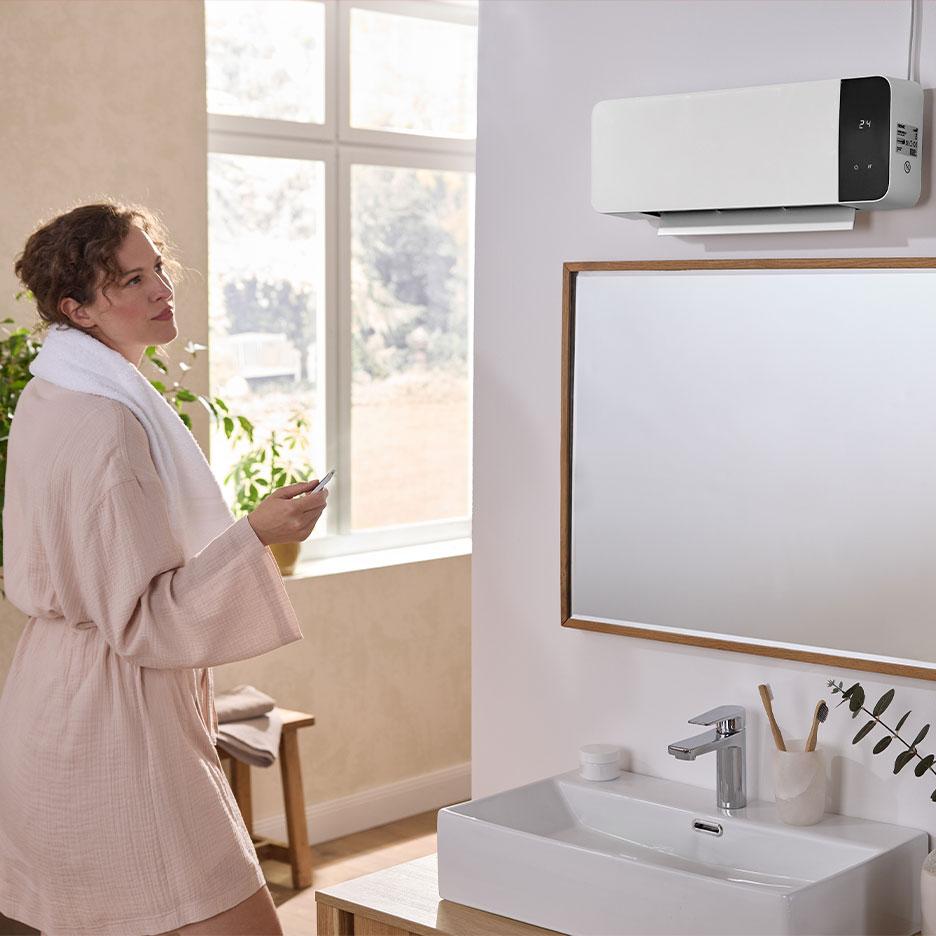 A woman in a bathrobe uses a remote control to operate a wall-mounted bathroom heater.