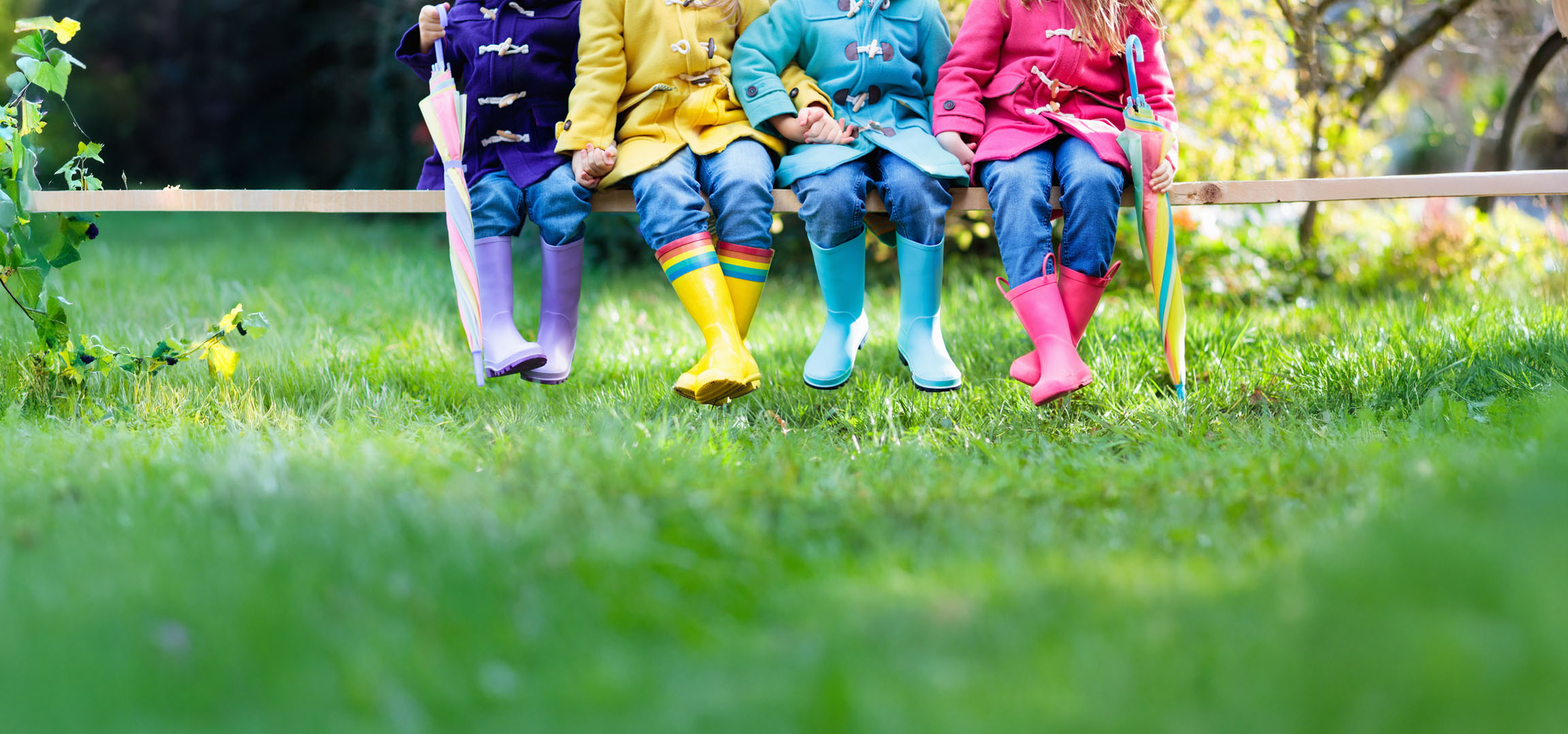 Children in colorful raincoats and boots sitting on a bench in the grass.