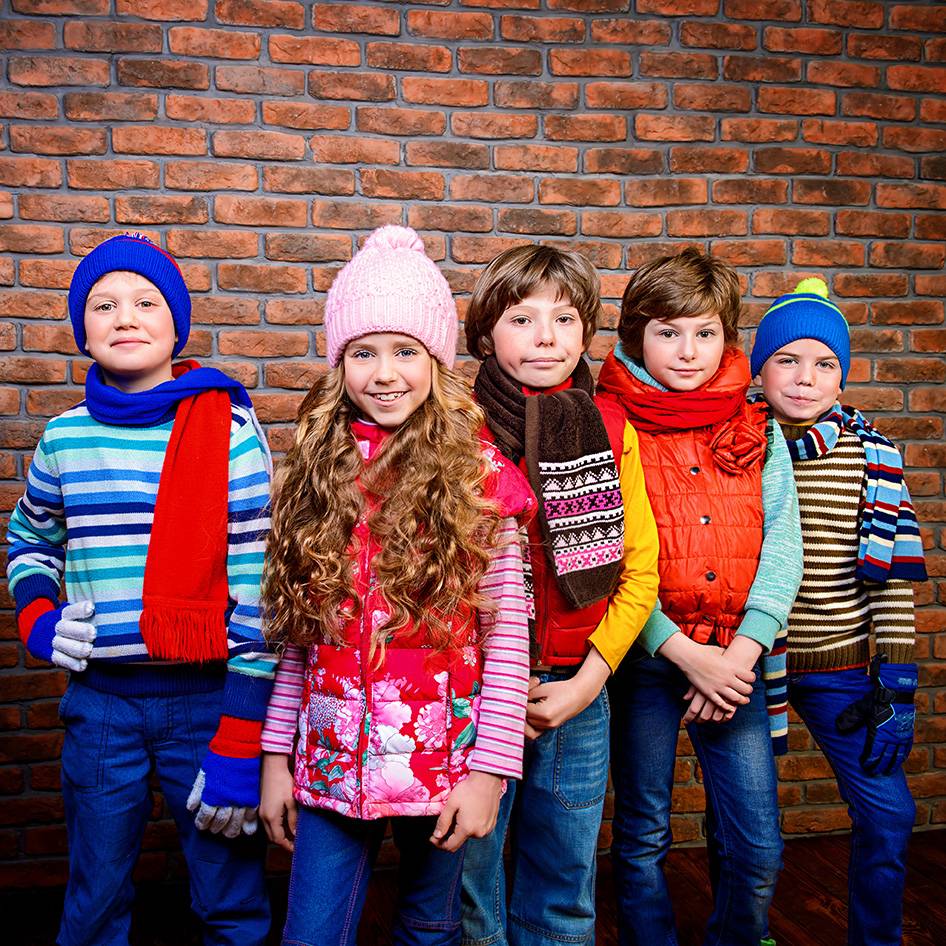 Five children in colorful winter hats, scarves, and vests against a brick wall.