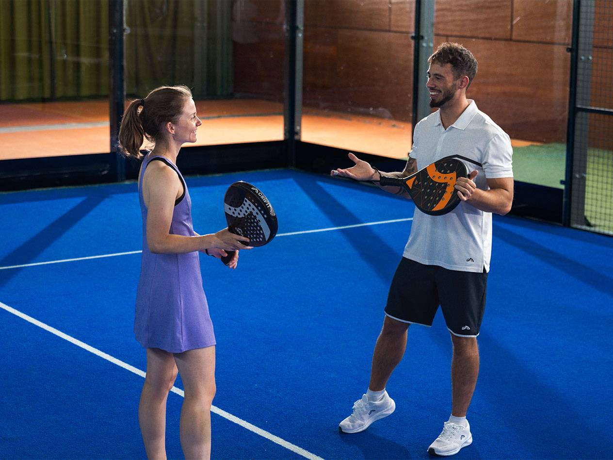 Two people on a padel court, holding padel rackets and conversing.