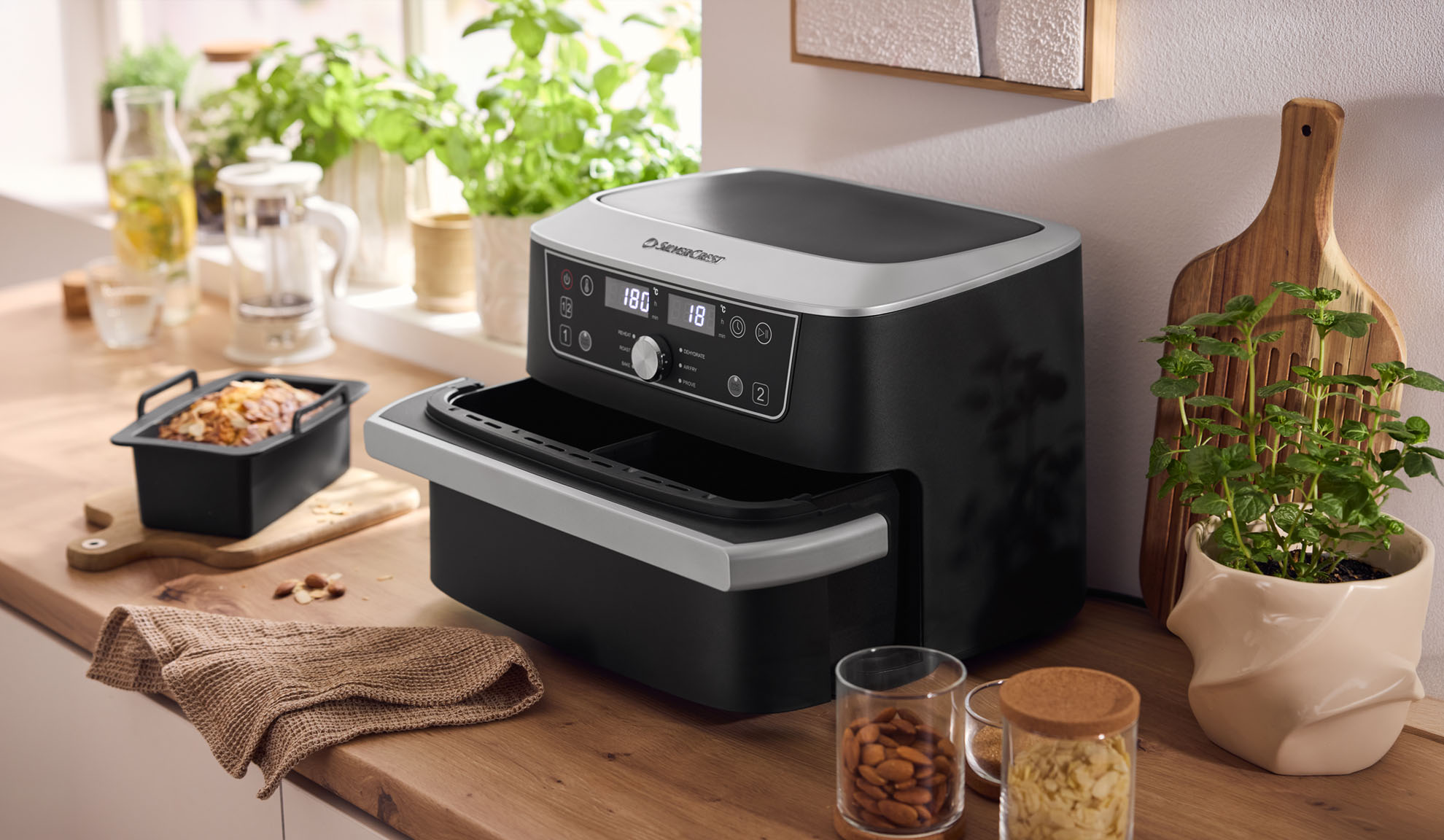 Black and silver air fryer on a wooden kitchen counter with baked goods and plants.