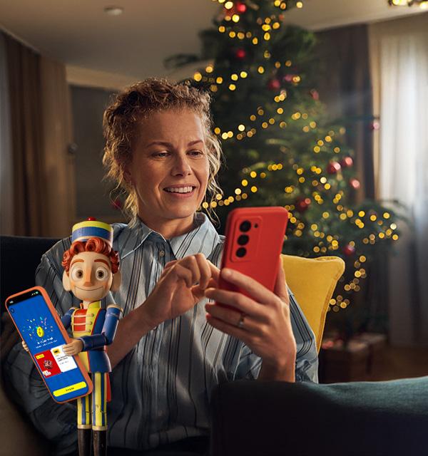 A smiling woman uses her phone next to a festive toy soldier in front of a Christmas tree.