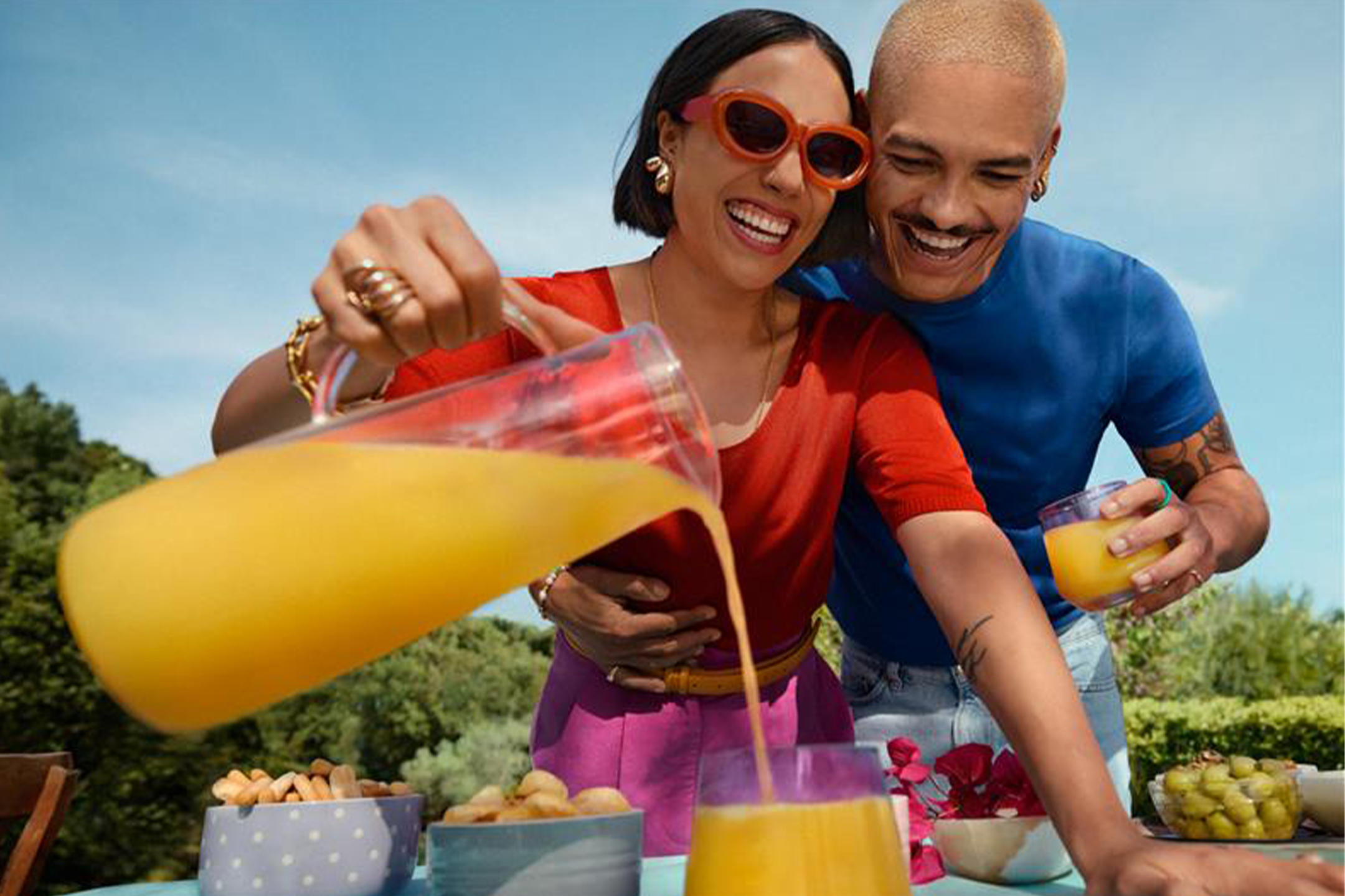 Two smiling people, one pouring a drink from a pitcher, against a clear blue sky.