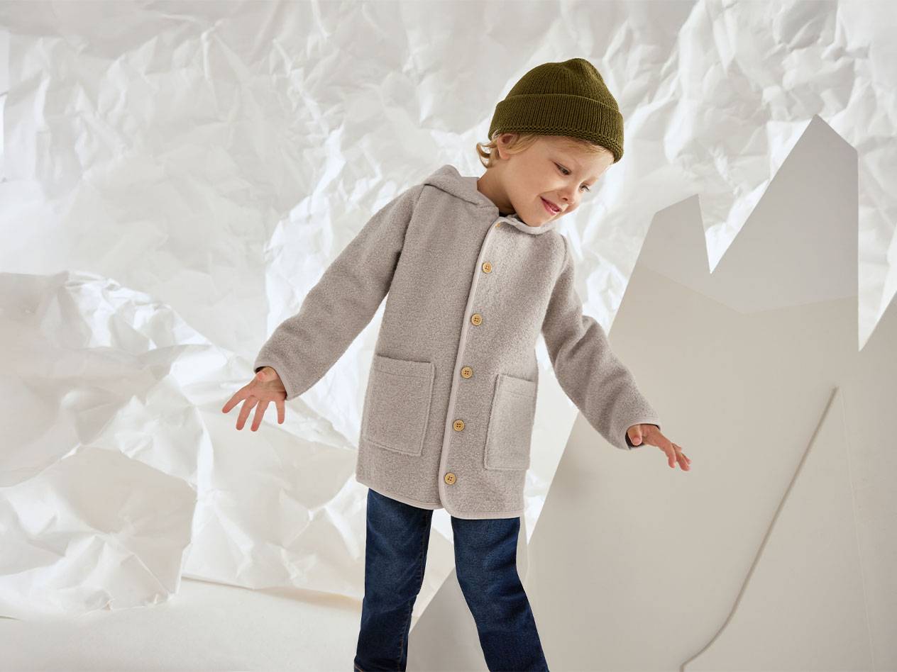 Boy in a grey wool jacket and green beanie, smiling against a crumpled paper background.