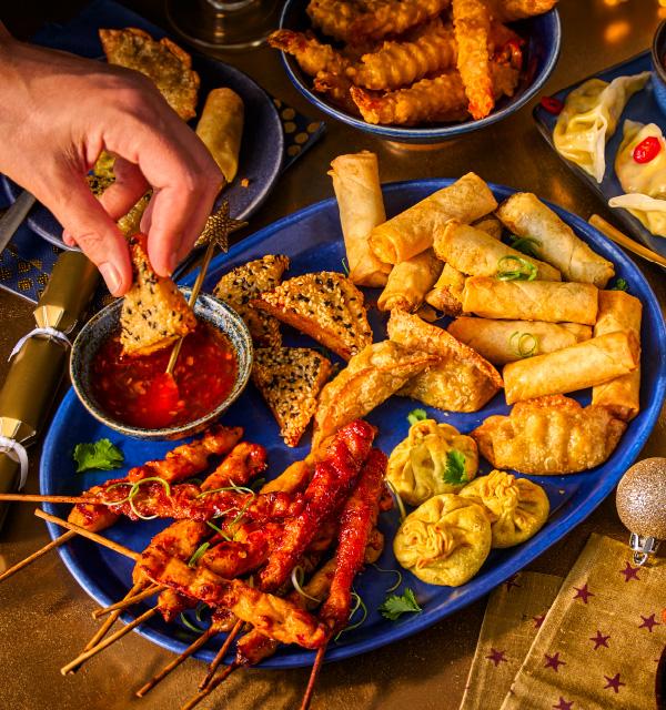 A festive platter of party food including spring rolls, chicken skewers, and dipping sauce.