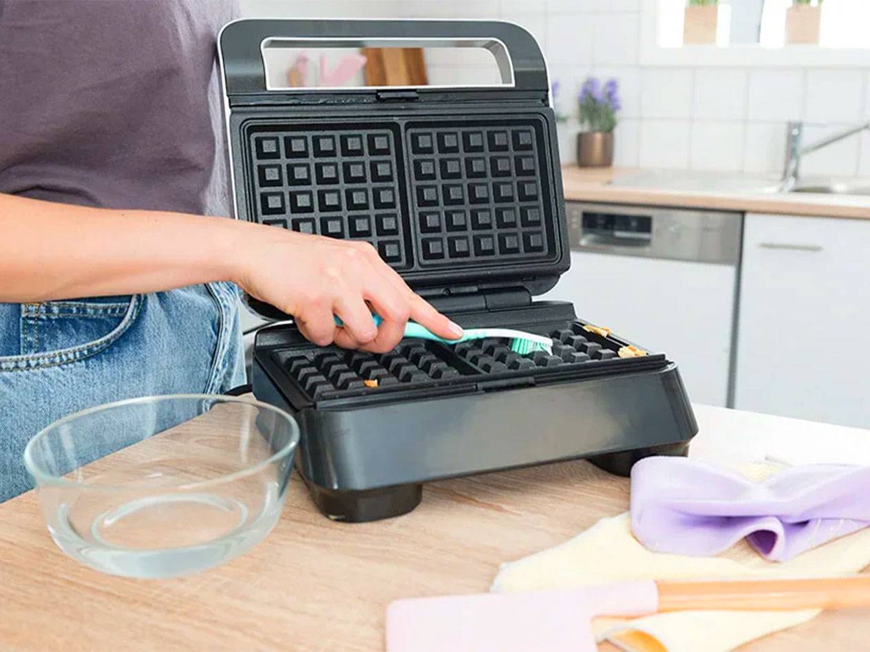 A person cleans a waffle maker with a toothbrush.