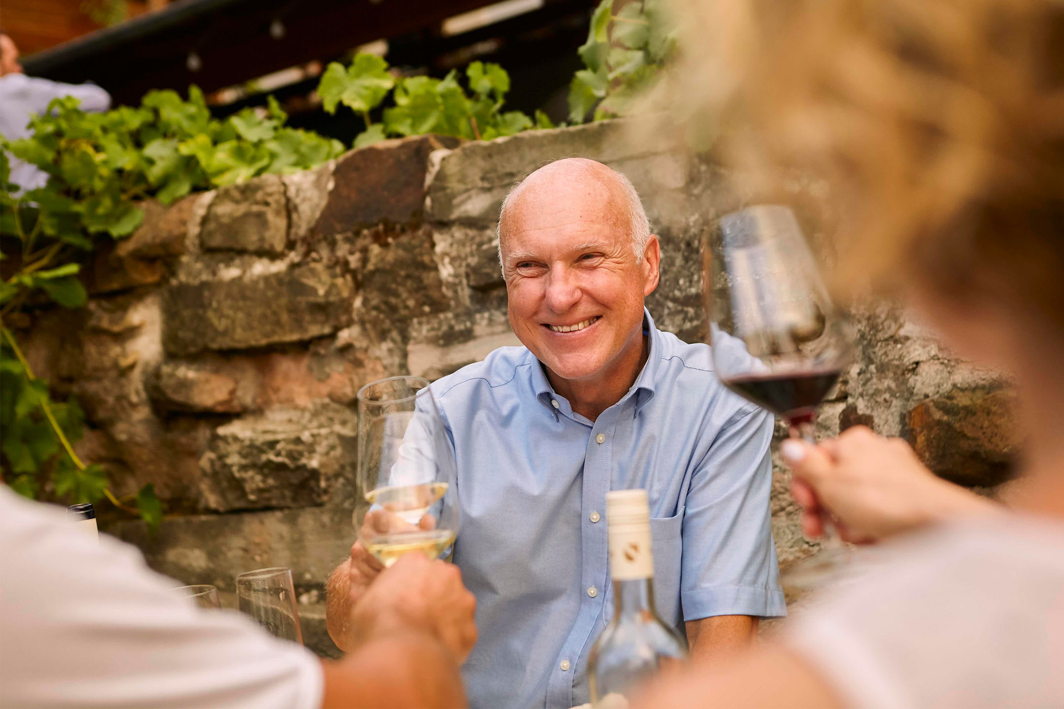 Smiling man toasting with white wine, surrounded by friends and a stone wall.