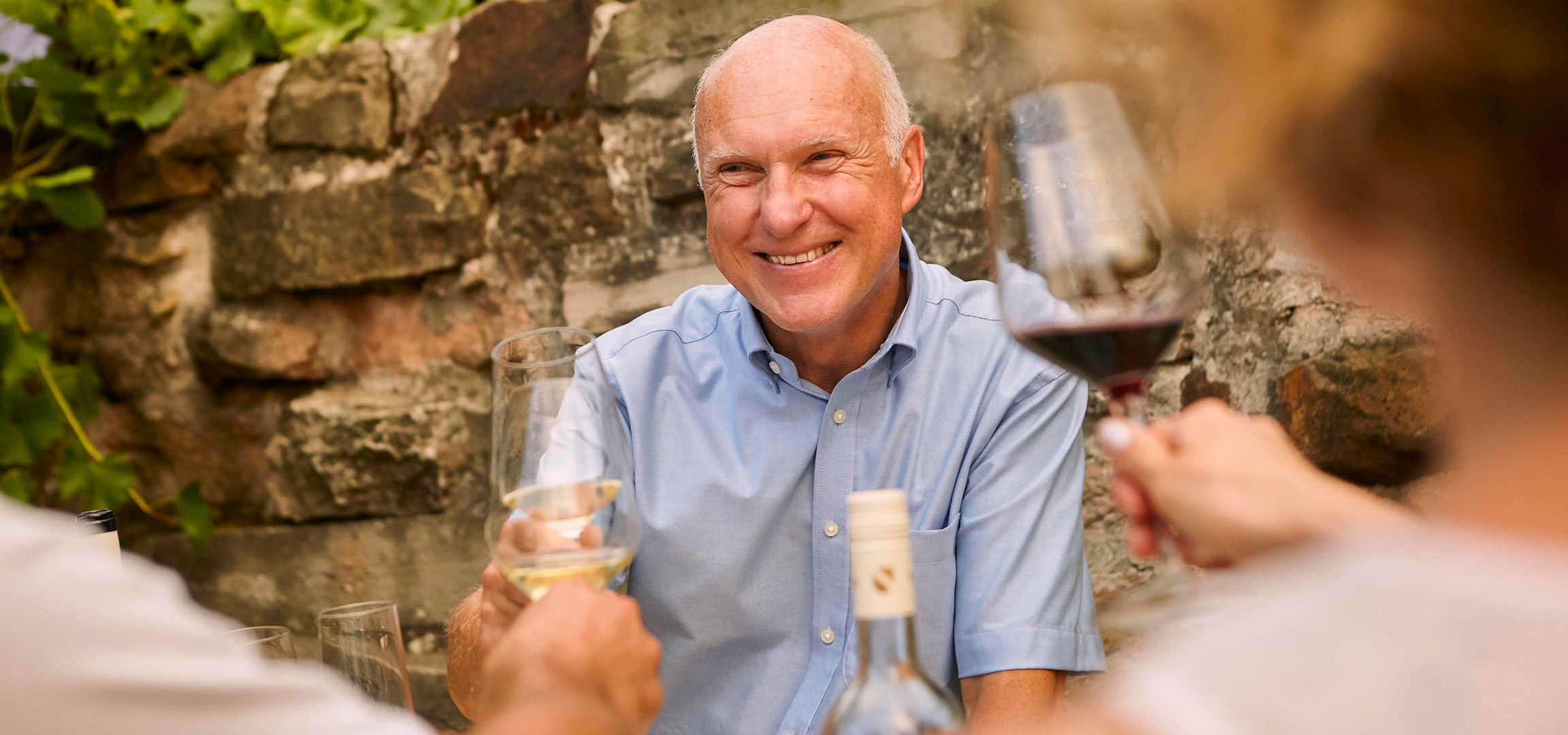 Man smiling with a glass of red wine on a terrace with a stone wall.