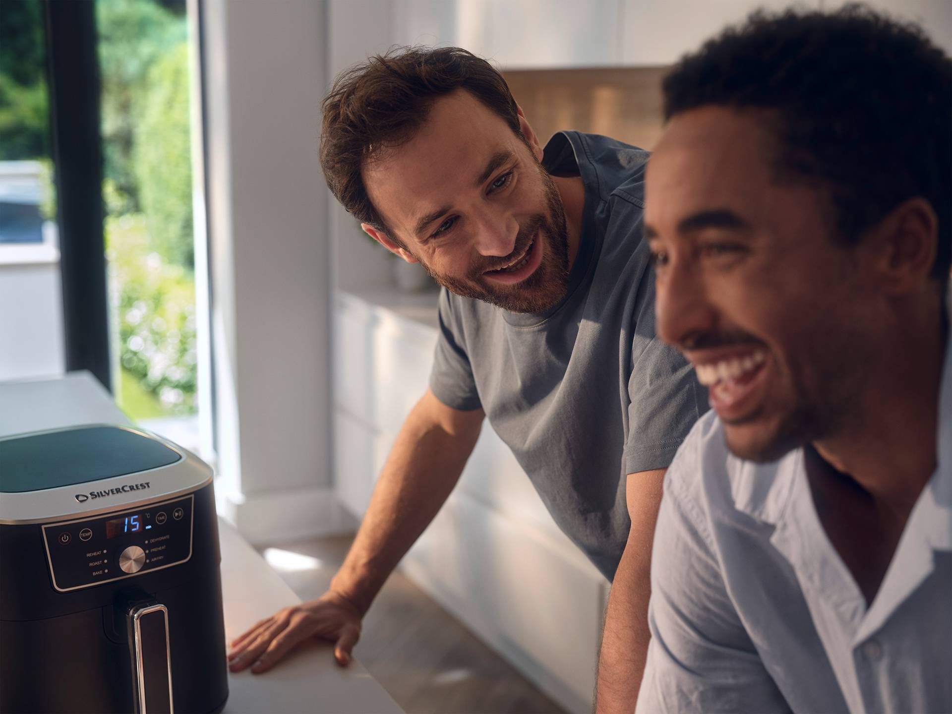 Two men smiling in a kitchen, with an air fryer on the counter showing '15' on its display.