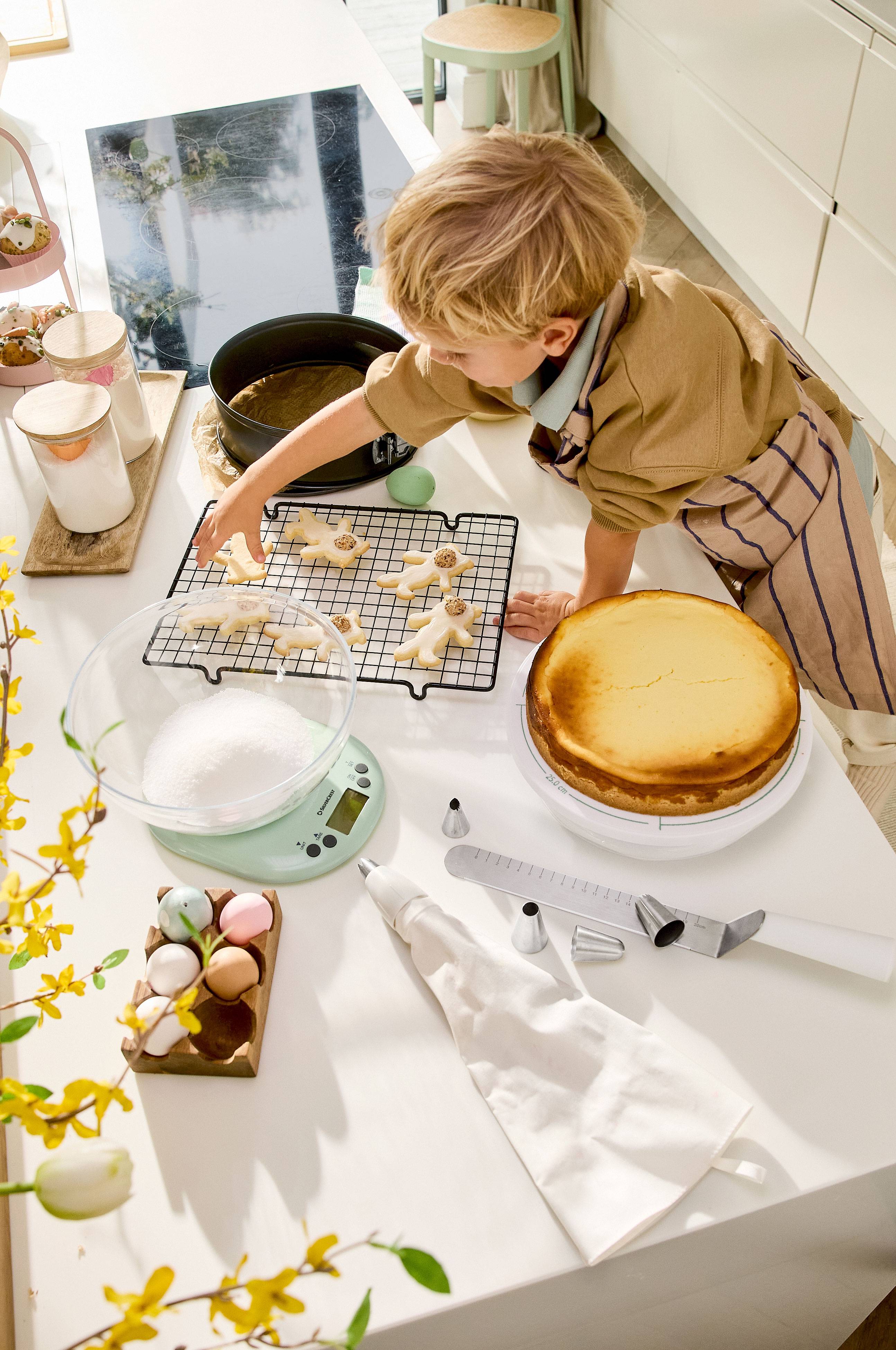 A child decorates Easter cookies, with a cheesecake and baking utensils on the table.