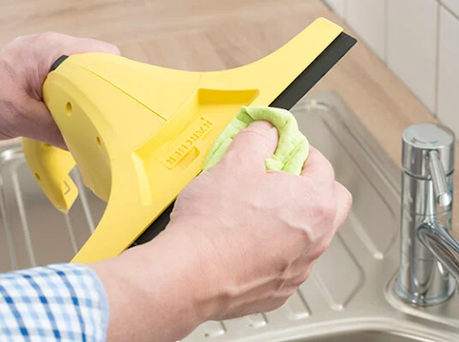 A person cleaning a yellow Kärcher window vacuum with a green cloth by a sink.