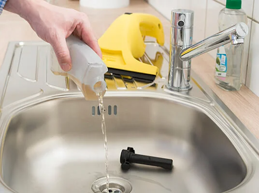 Hand pouring liquid from bottle into sink, with window cleaner and faucet in background.