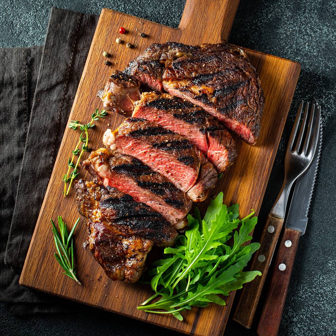 Sliced grilled steak on a wooden board with herbs and arugula.