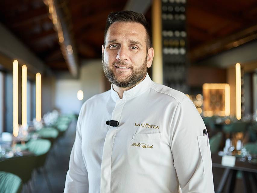 Smiling chef in white uniform with 'LA CONTEA' logo in an elegant restaurant.