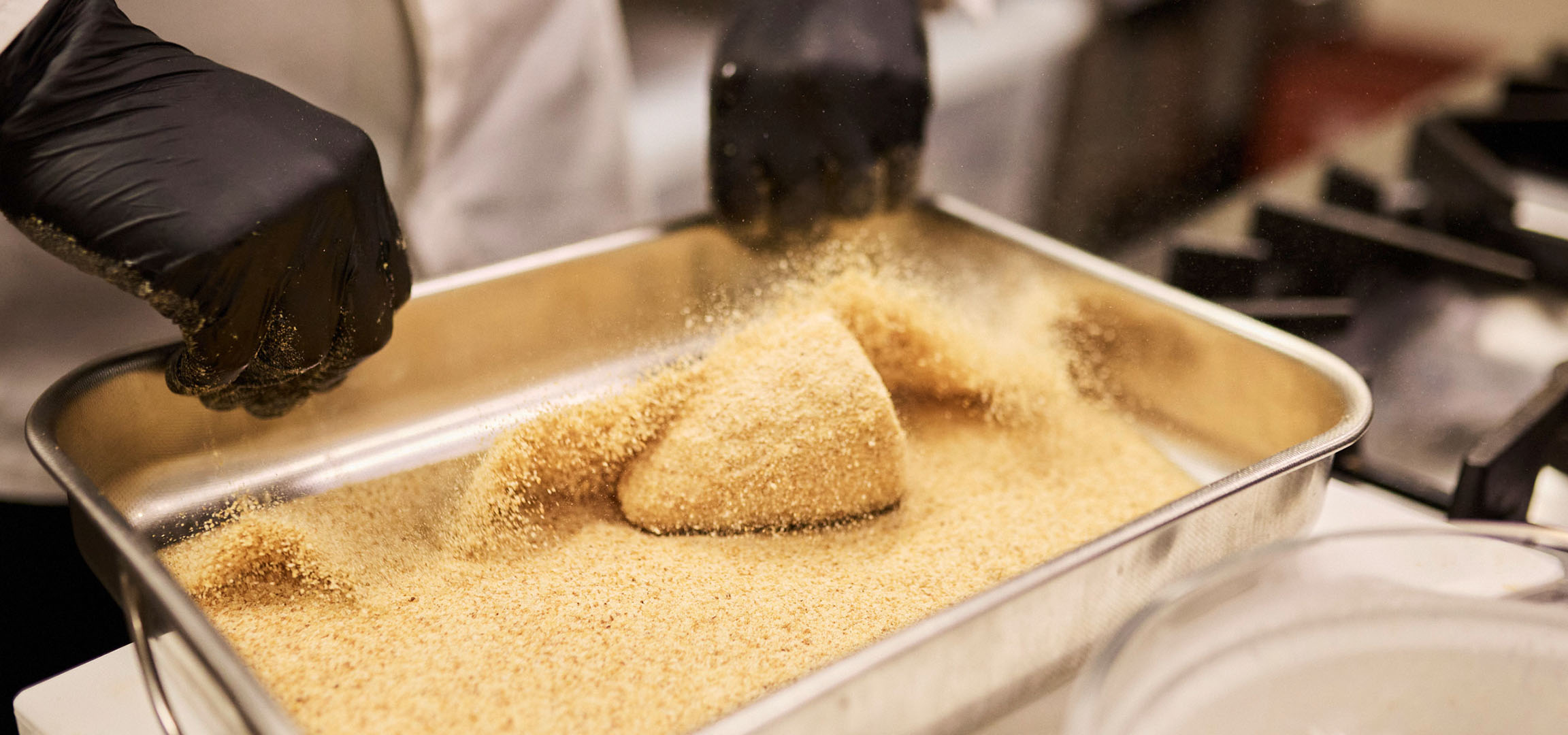 Chef breading food in breadcrumbs in a metal tray.