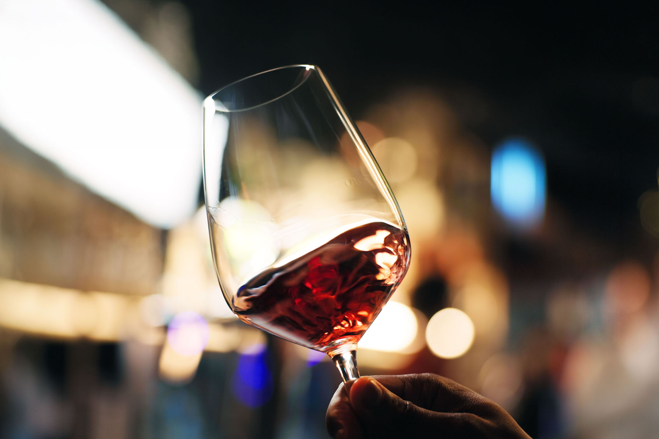 Glass of red wine held in hand, with blurred lights in the background.