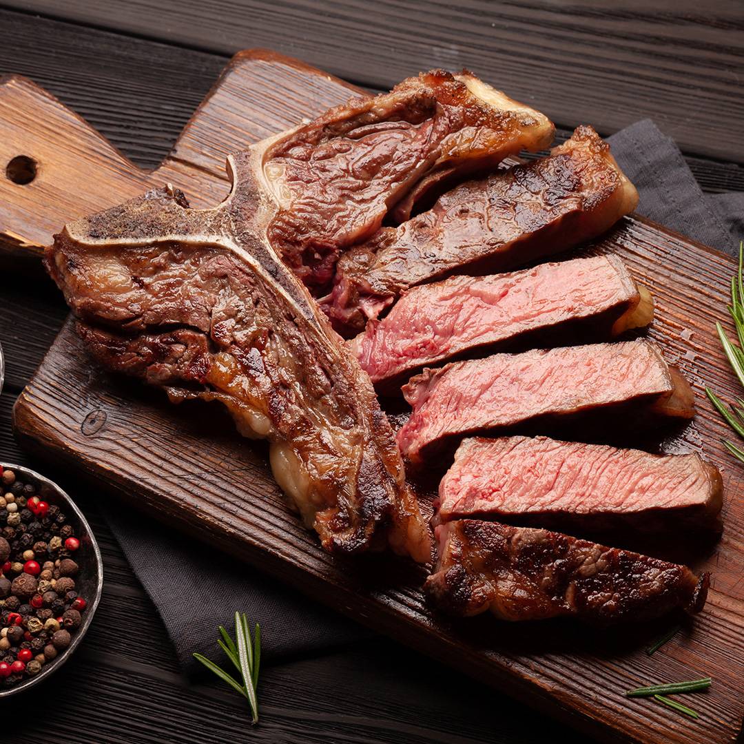 Cooked T-bone steak, sliced, on a wooden board with peppercorns and rosemary.