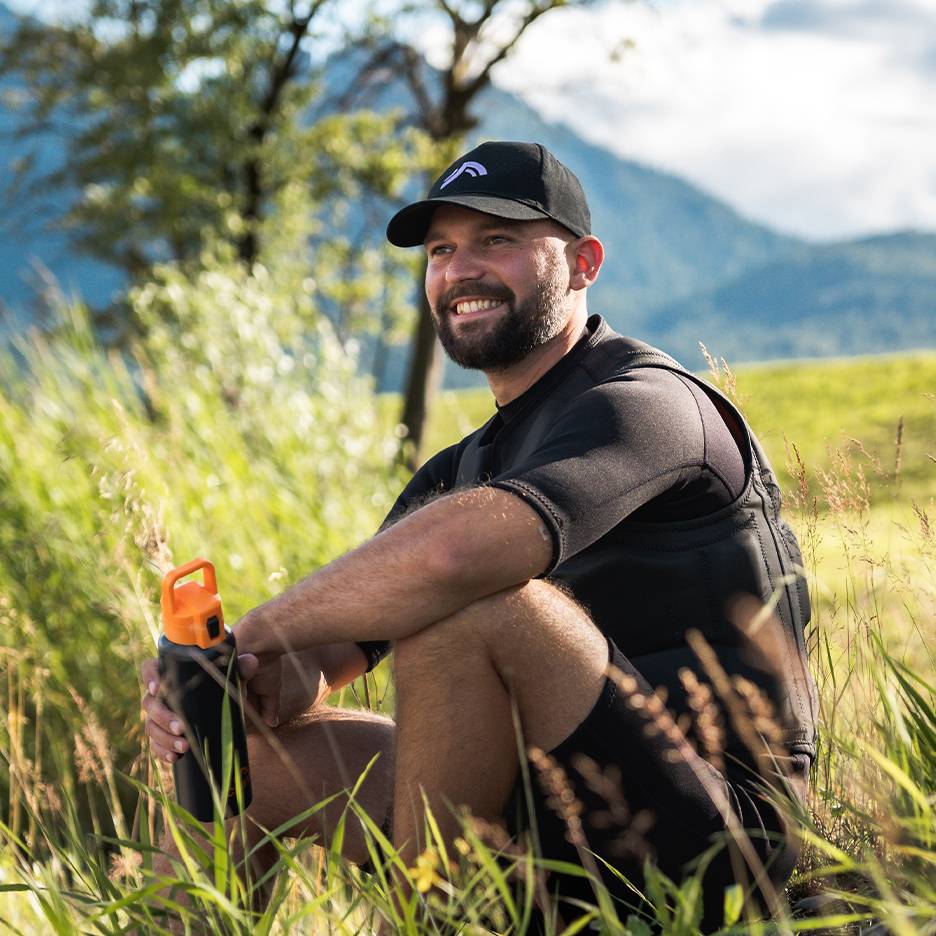 Smiling man in black cap and neoprene vest, holding an orange water bottle.