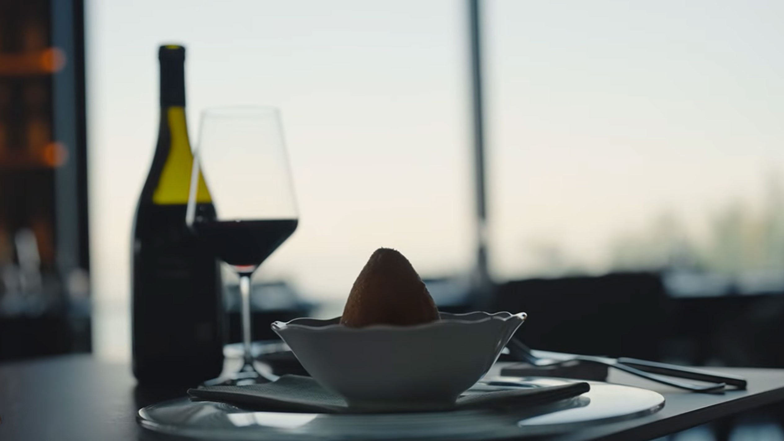 A bottle of red wine and a glass next to a single arancino in a bowl on a table.
